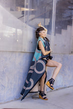 Load image into Gallery viewer, Woman holding a long, patterned scarf against a glass wall.