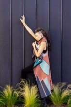 Load image into Gallery viewer, Person wearing a colorful dress standing against a dark blue wall with greenery at the base.