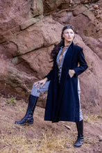 Load image into Gallery viewer, Person wearing a long dark coat standing against a rocky background