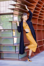 Load image into Gallery viewer, Woman in mustard yellow outfit and dark coat standing against a modern architectural wall.