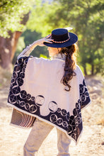 Load image into Gallery viewer, Woman wearing a black and white patterned shawl and blue hat outdoors.