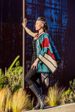 Load image into Gallery viewer, Woman wearing a colorful geometric-patterned poncho standing against a textured wall with plants in the foreground.