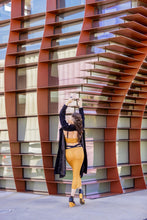 Load image into Gallery viewer, Person standing in front of a modern architectural structure with geometric design in backless black coat and gold dress.
