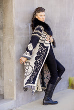 Load image into Gallery viewer, Person wearing a long, patterned coat with a fur-lined hood against a concrete wall.
