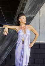 Load image into Gallery viewer, Woman in a light purple dress standing against a dark concrete wall.
