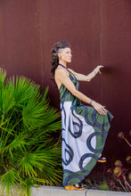 Load image into Gallery viewer, Woman in a patterned dress standing against a brown wall with greenery.

