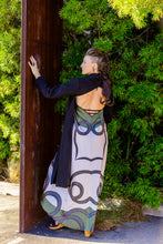Load image into Gallery viewer, Woman in a patterned dress standing against a wooden wall with greenery around
