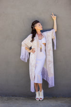 Load image into Gallery viewer, Woman wearing a long, light-colored fringe garment against a gray wall.
