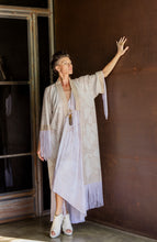 Load image into Gallery viewer, Woman in a long, light-colored robe with fringe standing against a dark wall.
