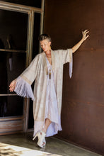 Load image into Gallery viewer, Woman in a long, light-colored kimono with fringe sleeves standing in a room with wooden floors and a dark wall.
