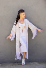 Load image into Gallery viewer, Woman wearing a long, light-colored fringe garment against a gray wall.
